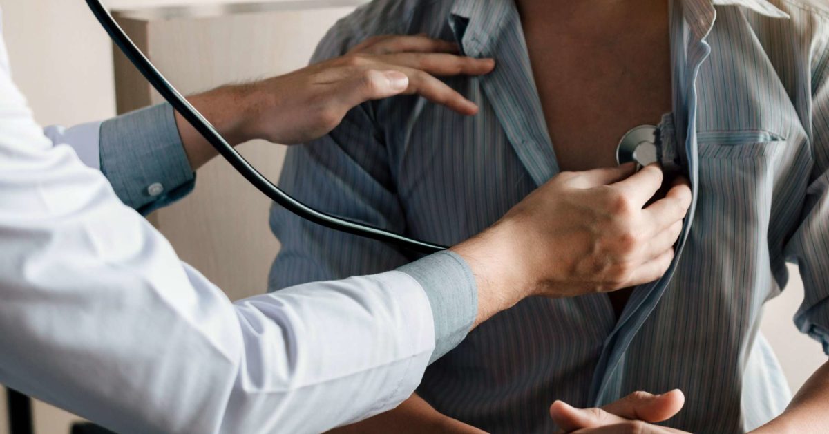  A doctor in a white coat uses a stethoscope to listen to a patient's heartbeat while another doctor stands nearby.